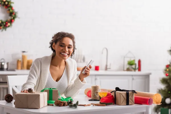 Donna afro-americana positiva che tiene il cellulare vicino ai regali, all'arredamento natalizio e al tè — Foto stock