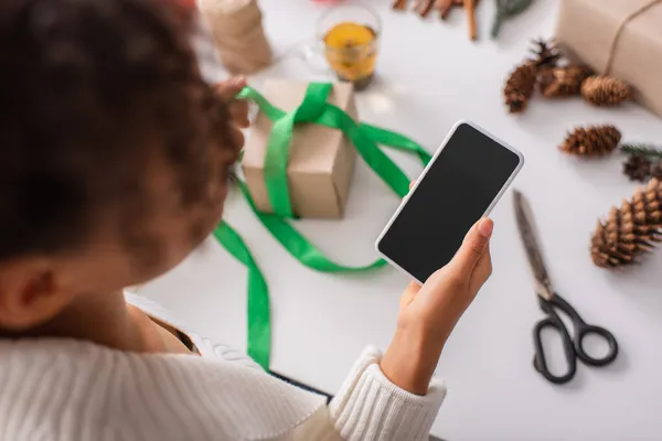 Vista aérea de una mujer afroamericana sosteniendo un teléfono celular cerca de regalos de navidad borrosos - foto de stock