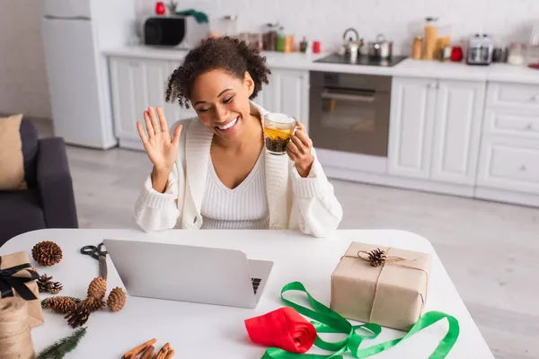 Heureuse femme afro-américaine avec thé ayant appel vidéo sur ordinateur portable près de cadeaux et décor de Noël — Photo de stock