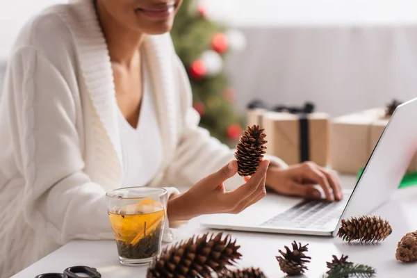 Cropped view of african american woman holding pine cone near tea and laptop at home — Stock Photo