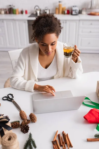 African american woman holding tea and using laptop near christmas decor at home — Stock Photo