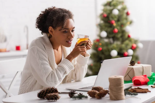 African american woman holding tea near laptop, present and christmas decor — Stock Photo
