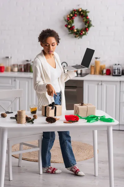 African american woman with laptop pointing at gift near christmas decor at home — Stock Photo