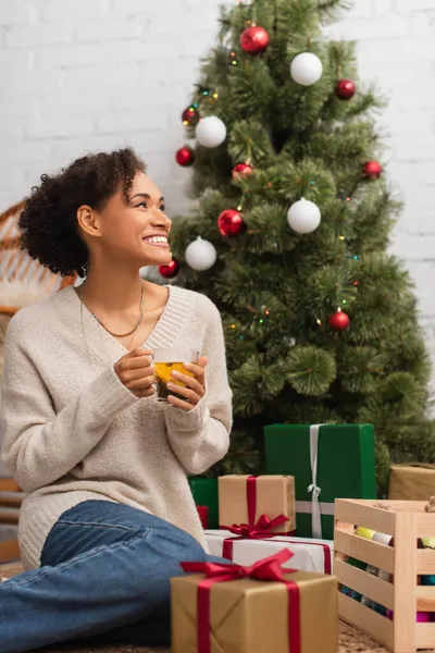 Cheerful african american woman holding tea near presents and christmas tree — Stock Photo