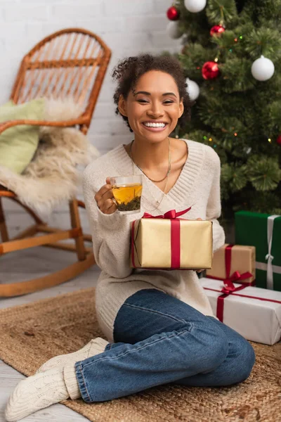 Positive african american woman holding gift and cup of tea near christmas tree at home — Stock Photo