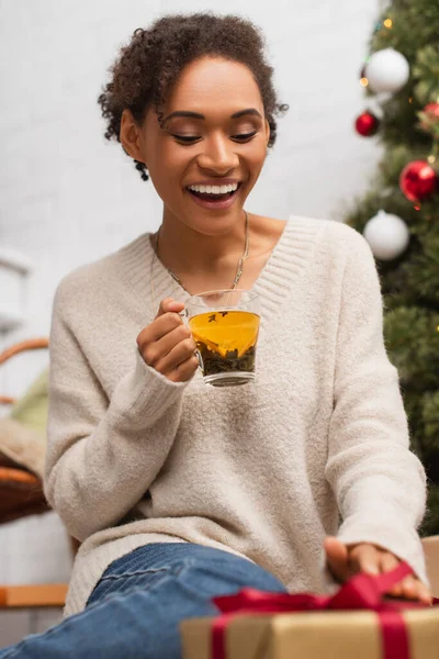 Happy african american woman with cup of tea looking at blurred christmas gift at home — Stock Photo