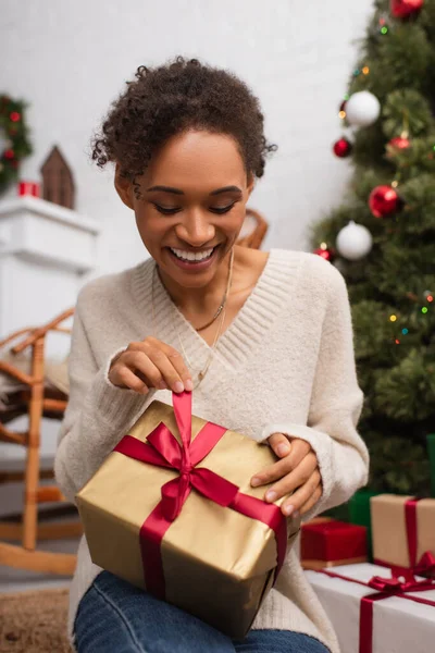Joven mujer afroamericana sonriendo mientras desata el regalo de Navidad en casa - foto de stock