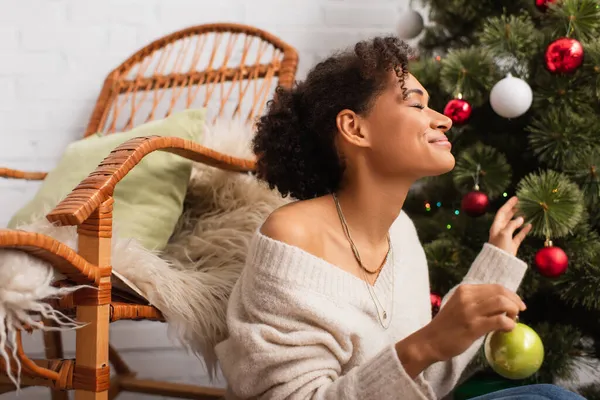 Vista lateral da mulher americana africana alegre segurando bola de Natal perto de árvore e cadeira em casa — Fotografia de Stock