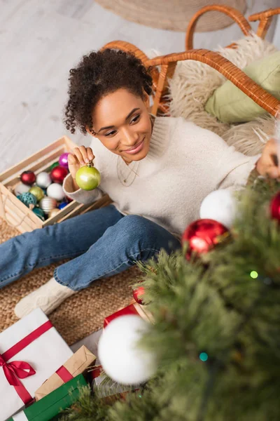 Vista de ângulo alto de mulher americana africana sorridente segurando bola perto de árvore de Natal borrada e presentes em casa — Fotografia de Stock