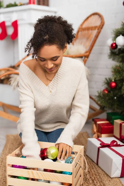 African american woman looking at box with christmas balls near gifts at home — Stock Photo