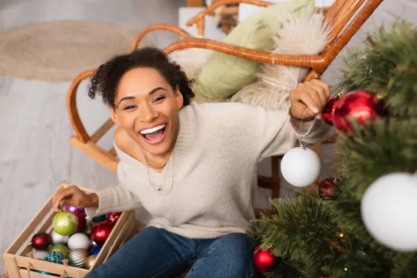 Vista ad alto angolo di donna afro-americana eccitata guardando la fotocamera mentre decorava l'albero di Natale a casa — Foto stock