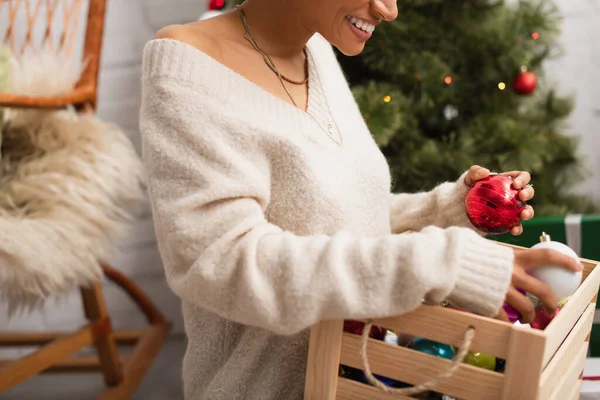 Vue recadrée d'une femme afro-américaine souriante tenant des boules de Noël dans une boîte à la maison — Photo de stock