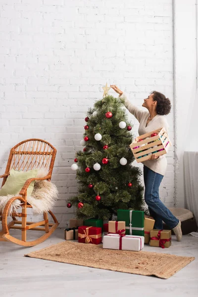 Vista laterale della donna afro-americana positiva decorare l'albero di Natale vicino ai regali a casa — Foto stock