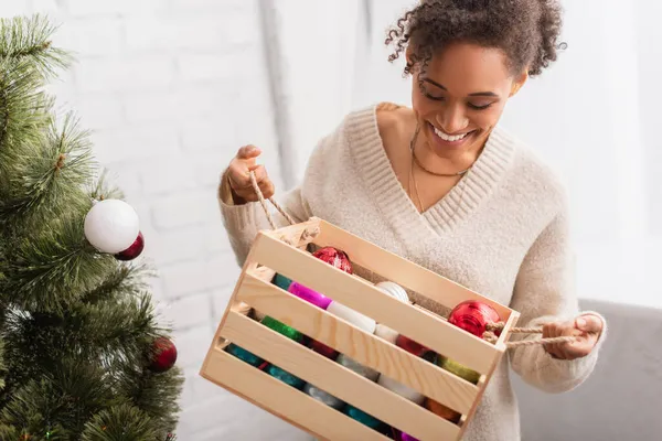 Smiling african american woman holding wooden box with balls near christmas tree at home — Stock Photo