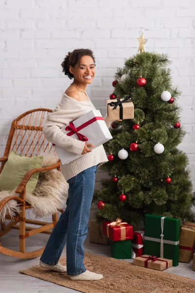 Sonriente mujer afroamericana mirando a la cámara mientras sostiene regalos cerca del árbol de Navidad en casa - foto de stock