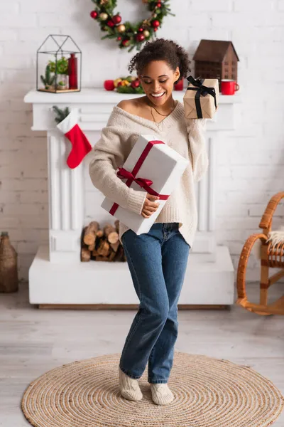 Cheerful african american woman holding presents near fireplace and christmas wreath — Stock Photo