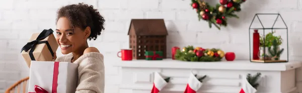 Afroamerikanerin hält Weihnachtsgeschenke neben dekoriertem Kamin zu Hause, Banner — Stockfoto