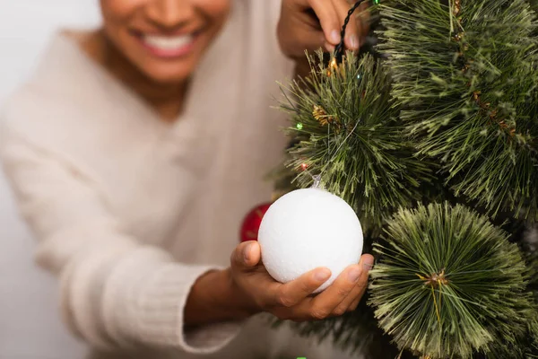 Vista recortada de la borrosa mujer afroamericana sosteniendo la bola de Navidad en el pino en casa - foto de stock