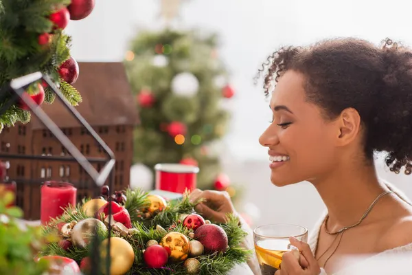 Side view of smiling african american woman holding cup of tea near decorated christmas wreath — Stock Photo