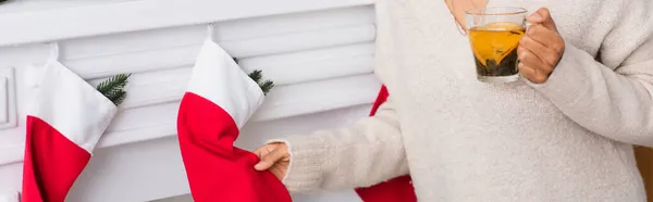 Cropped view of african american woman with tea holding christmas stocking on fireplace, banner — Stock Photo
