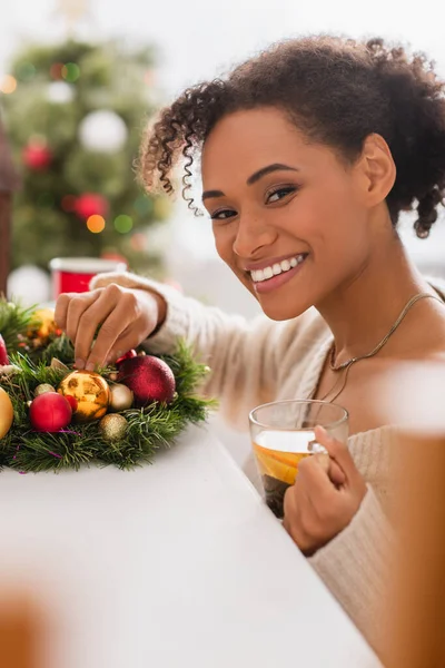 Fröhliche afrikanisch-amerikanische Frau mit Tee blickt in die Kamera in der Nähe des Weihnachtskranzes — Stockfoto