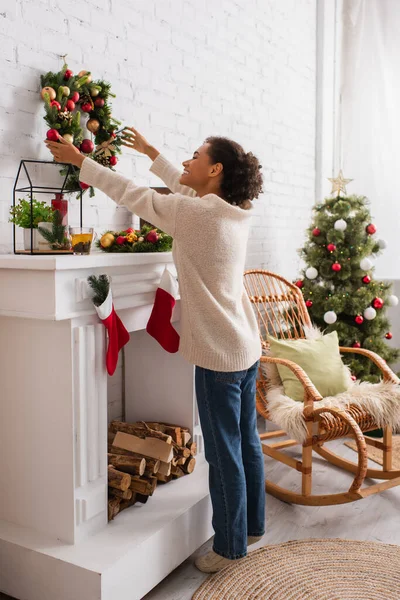 Vue latérale d'une femme afro-américaine positive tenant une couronne de Noël près d'une cheminée décorée à la maison — Photo de stock
