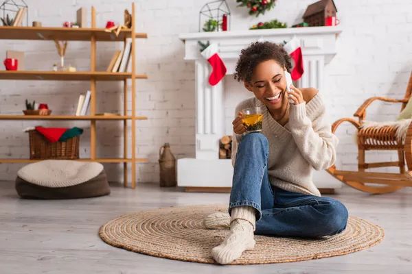 Femme afro-américaine souriante avec tasse de thé parlant sur un téléphone portable près d'une cheminée décorée à la maison — Photo de stock