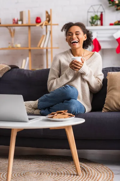 Riéndose mujer afroamericana sosteniendo taza cerca de la computadora portátil y galletas de Navidad en casa - foto de stock