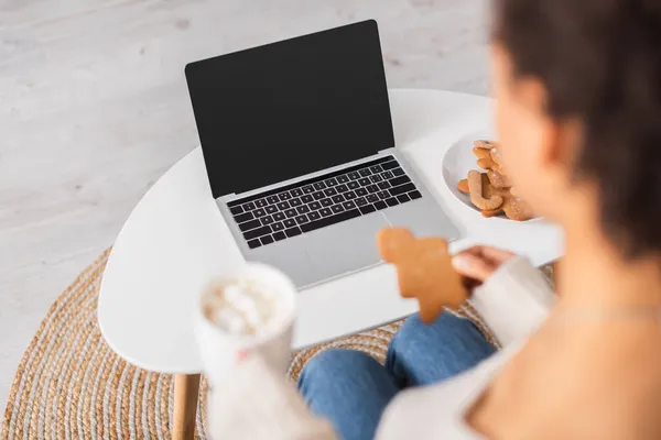 Mujer afroamericana borrosa sosteniendo la galleta de Navidad y la taza cerca de la computadora portátil en casa - foto de stock