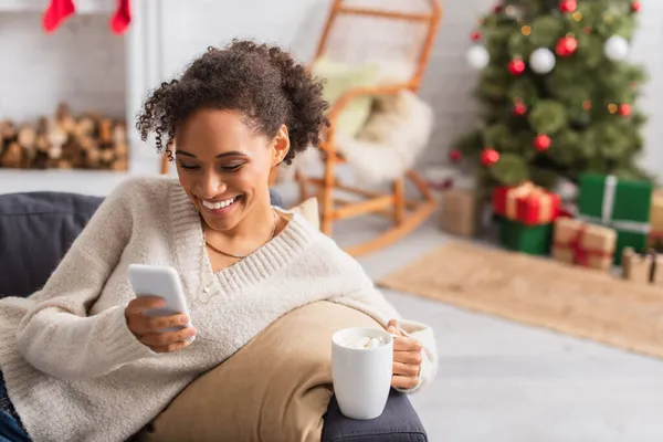 Mulher americana africana alegre segurando smartphone e xícara de cacau com marshmallows em casa — Fotografia de Stock