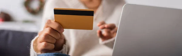 Vista recortada de la tarjeta de crédito en la mano de la mujer afroamericana borrosa cerca de la computadora portátil en casa, bandera - foto de stock