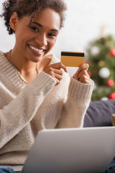 Улыбающаяся американка из Африки держит кредитку возле размытого ноутбука во время Рождества дома — стоковое фото