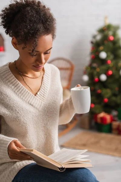Afroamerikanerin hält Tasse in der Hand, während sie zu Weihnachten zu Hause Buch liest — Stockfoto