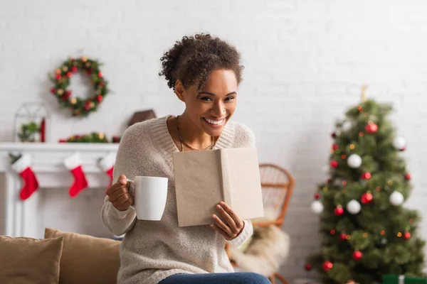 Femme afro-américaine souriante avec tasse et livre regardant la caméra pendant Noël à la maison — Photo de stock