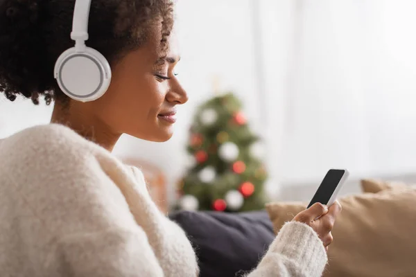 Side view of african american woman in headphones using cellphone during christmas at home — Stock Photo