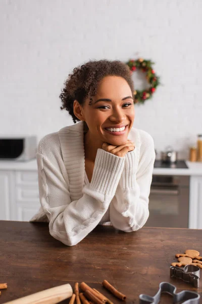 Cheerful african american woman looking at camera near cinnamon sticks and cookie cutters in kitchen — Stock Photo