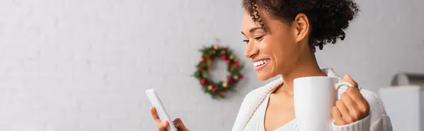 Junge afrikanisch-amerikanische Frau mit Tasse mit Smartphone während Weihnachten zu Hause, Banner — Stockfoto