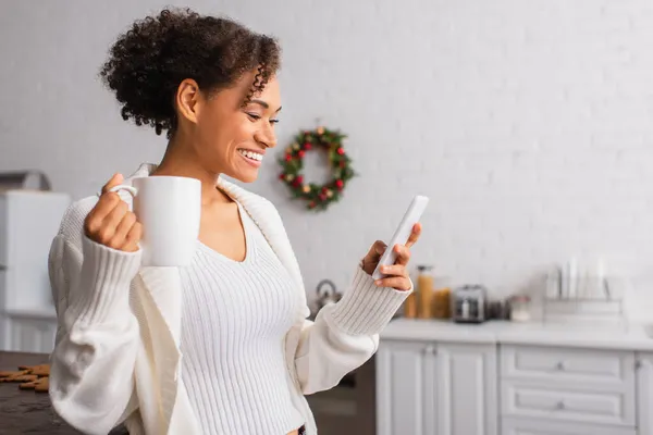 Sonriente mujer afroamericana con taza usando teléfono móvil en la cocina durante la Navidad - foto de stock
