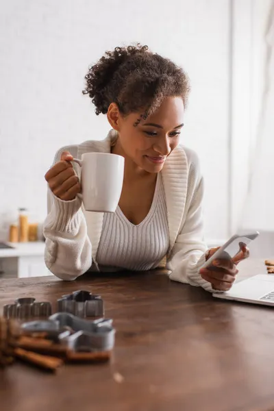 Mujer afroamericana con taza usando smartphone cerca de computadora portátil y cortadores de galletas en la cocina - foto de stock