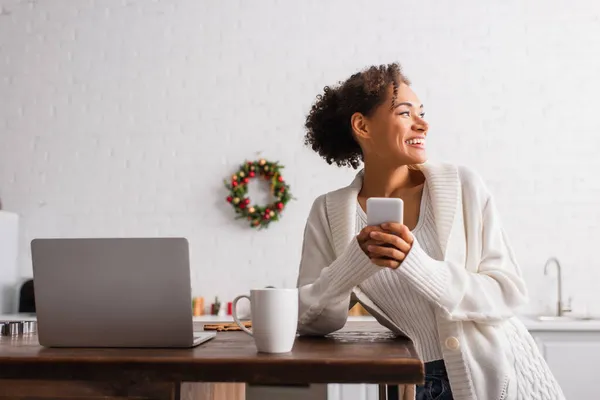 Positive african american woman using smartphone near cup and laptop in kitchen on christmas — Stock Photo
