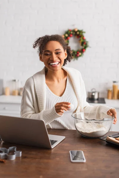 Femme afro-américaine joyeuse tenant un oeuf près de la farine et des appareils à la maison — Photo de stock