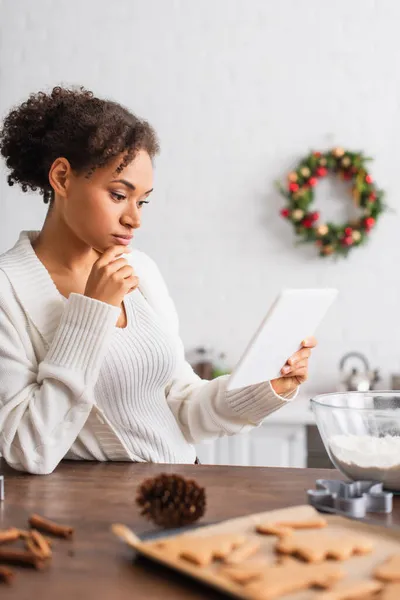 Young african american woman using digital tablet near blurred gingerbreads and flour in kitchen — Stock Photo
