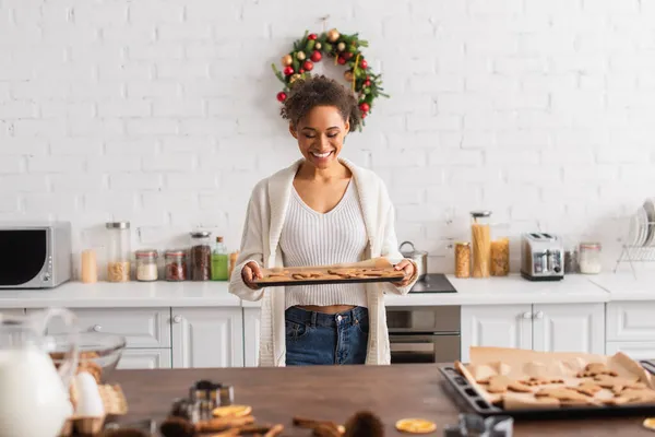 Sorridente donna africana americana che tiene pan di zenzero al forno vicino agli ingredienti in cucina — Foto stock