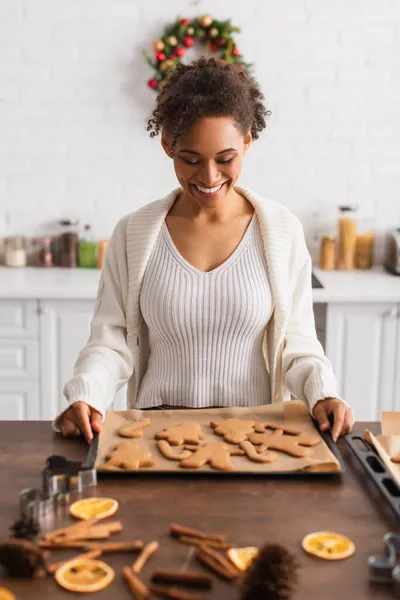 Happy african american woman holding tray with christmas gingerbreads near cinnamon sticks — Stock Photo