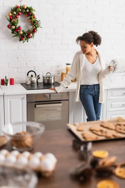 African american woman with smartphone standing near stove and christmas wreath in kitchen — Stock Photo