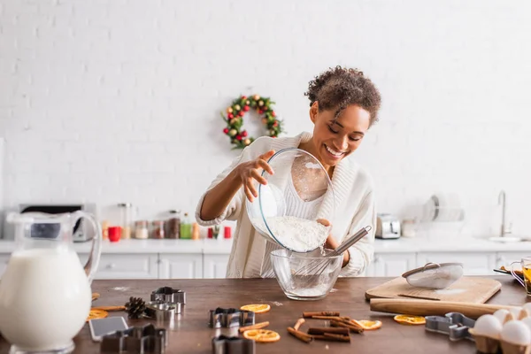 Mulher afro-americana positiva derramando farinha enquanto cozinha perto de cortadores de biscoitos na cozinha — Fotografia de Stock