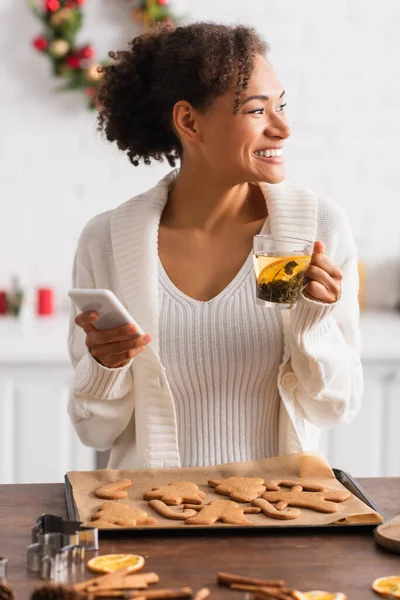 Улыбающаяся африканская американка с чашкой чая и смартфоном возле запеченного рождественского пряника — стоковое фото