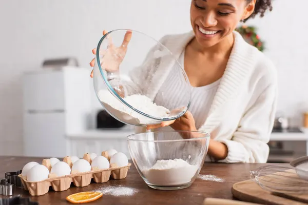 Sorrindo mulher afro-americana derramando farinha perto de ovos e cortadores de biscoitos na cozinha — Fotografia de Stock