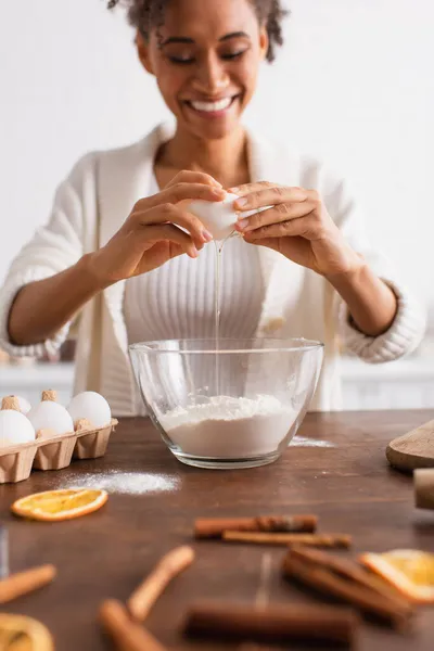 Donna afroamericana offuscata che versa l'uovo nella farina vicino ai bastoncini di cannella in cucina — Foto stock