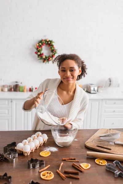 Mujer afroamericana sonriente vertiendo harina cerca de ingredientes, palitos de canela y naranja seca en la cocina - foto de stock